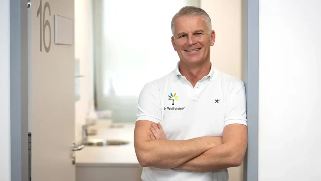 Profil Dr. Jochen Wolfsteiner Dr. Jochen Wolfsteiner lehnt in der Tür zum Behandlungszimmer mit verschränkten Armen und lächelt