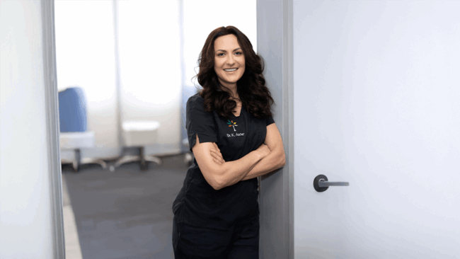 Profil Dr. Kornelia Ascher Dr. Kornelia Ascher lehnt in der Tür zum Behandlungszimmer mit verschränkten Armen und lächelt