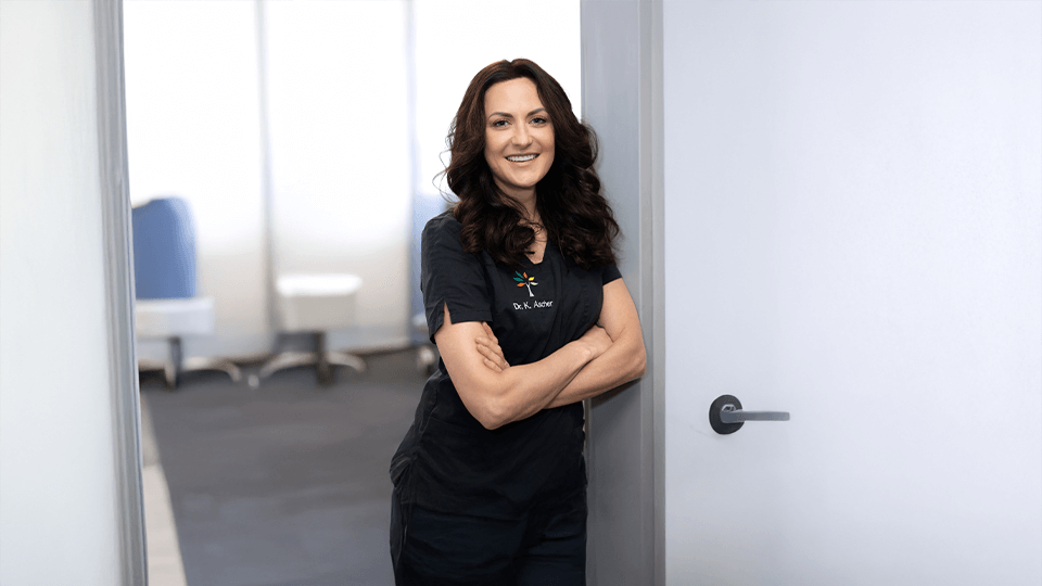 Dr. Kornelia Ascher lehnt in der Tür zum Behandlungszimmer mit verschränkten Armen und lächelt