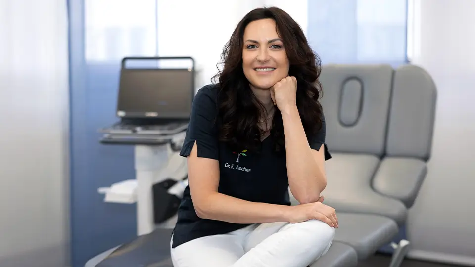 Dr. Kornelia Ascher sitzt auf einer Behnaldungsliege und stützt Ihrem Kopf auf der Hand ab