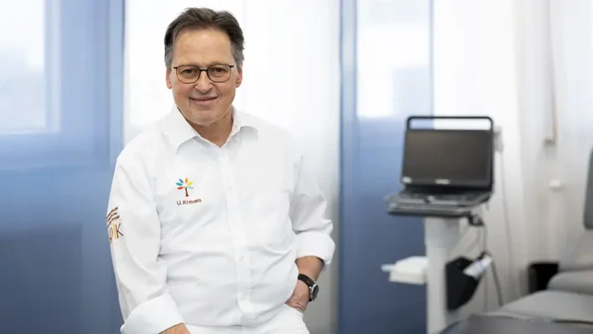Profil Ulrich Kreuels Ulrich Kreuels – Facharzt für Orthopädie trägt einen weißen Kittel mit Logo und sitzt in seiner hellen, modernen Praxis