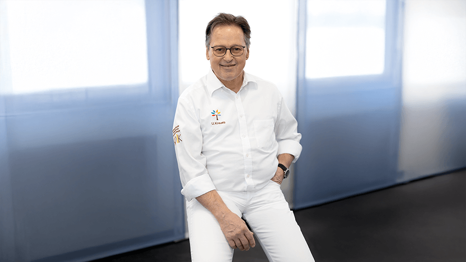 Ulrich Kreuels – Facharzt für Orthopädie trägt einen weißen Kittel mit Logo und sitzt in seiner hellen, modernen Praxis