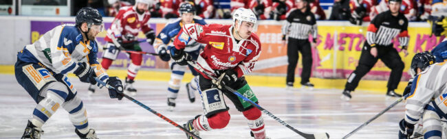 Eishockeyspieler in Aktion auf dem Eis, zwei Spieler in rot-weißer und zwei in blau-weißer Ausrüstung kämpfen um den Puck, im Hintergrund Schiedsrichter und Zuschauer.