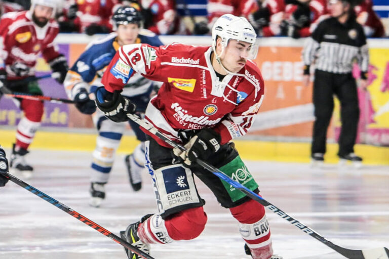 Eishockeyspieler in Aktion auf dem Eis, zwei Spieler in rot-weißer und zwei in blau-weißer Ausrüstung kämpfen um den Puck, im Hintergrund Schiedsrichter und Zuschauer.